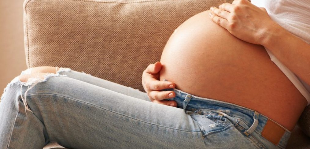 A pregnant woman sitting down on a tan couch with her hands around her stomach.