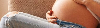 A pregnant woman sitting down on a tan couch with her hands around her stomach.