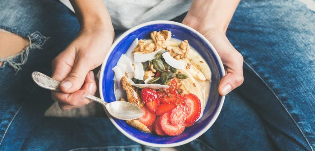 Yogurt, granola, seeds, fresh and dry fruits and honey in blue ceramic bowl in woman' s hands.