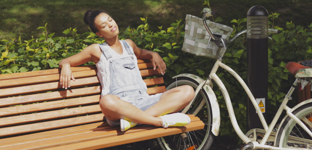 Young casual woman sitting with legs crossed and eyes closed on wooden bench in park with bicycle near and enjoying sunlight.