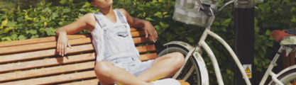 Young casual woman sitting with legs crossed and eyes closed on wooden bench in park with bicycle near and enjoying sunlight.