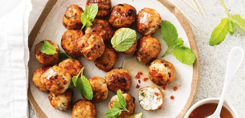 meatballs on a plate with fresh herbs and a bowl of chilli sauce with a spoon in it